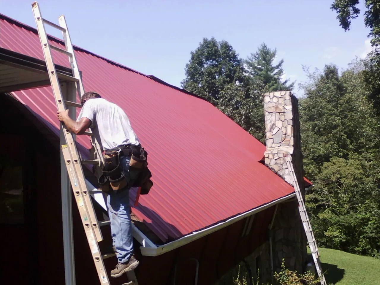 Roofer on a ladder beside a red metal roof and stone chimney — Soffit & Fascia Repair by Roofing Brothers in Mullens